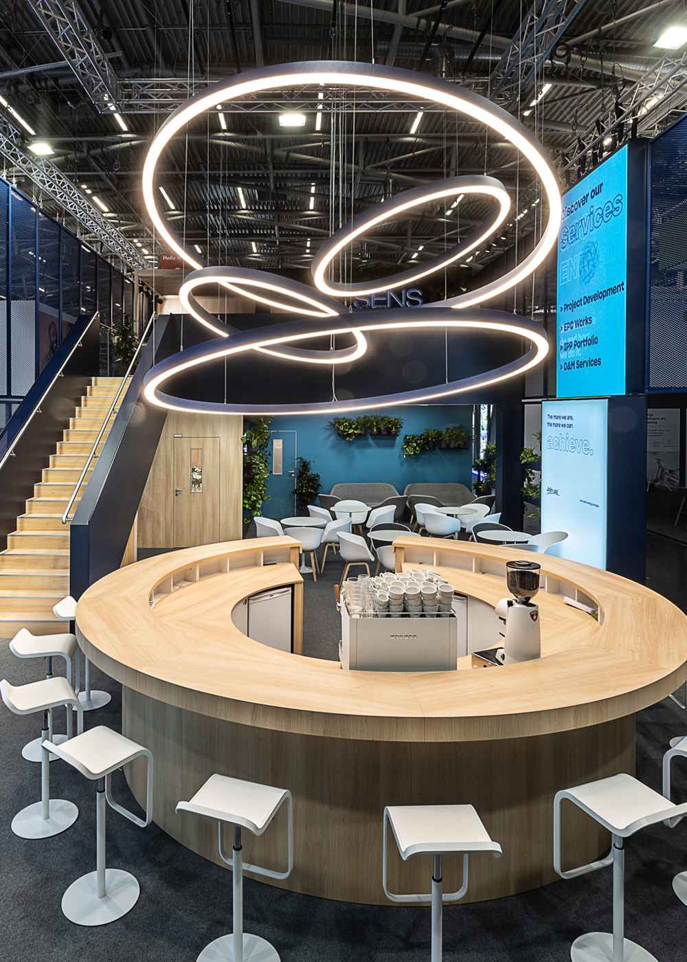 Zentrale Kommunikationsbar des zweigeschossigen SENS Messestands auf der Intersolar Smarter E in München mit organisch geformter Holztheke, markantem LED-Ringlicht als Blickfang, offenen Sitzbereichen, begrünten Loungezonen und transparenter Streckmetall-Architektur. Das einladende Standkonzept verbindet Design, Nachhaltigkeit und Kommunikation auf einer 192 m² großen Ausstellungsfläche.