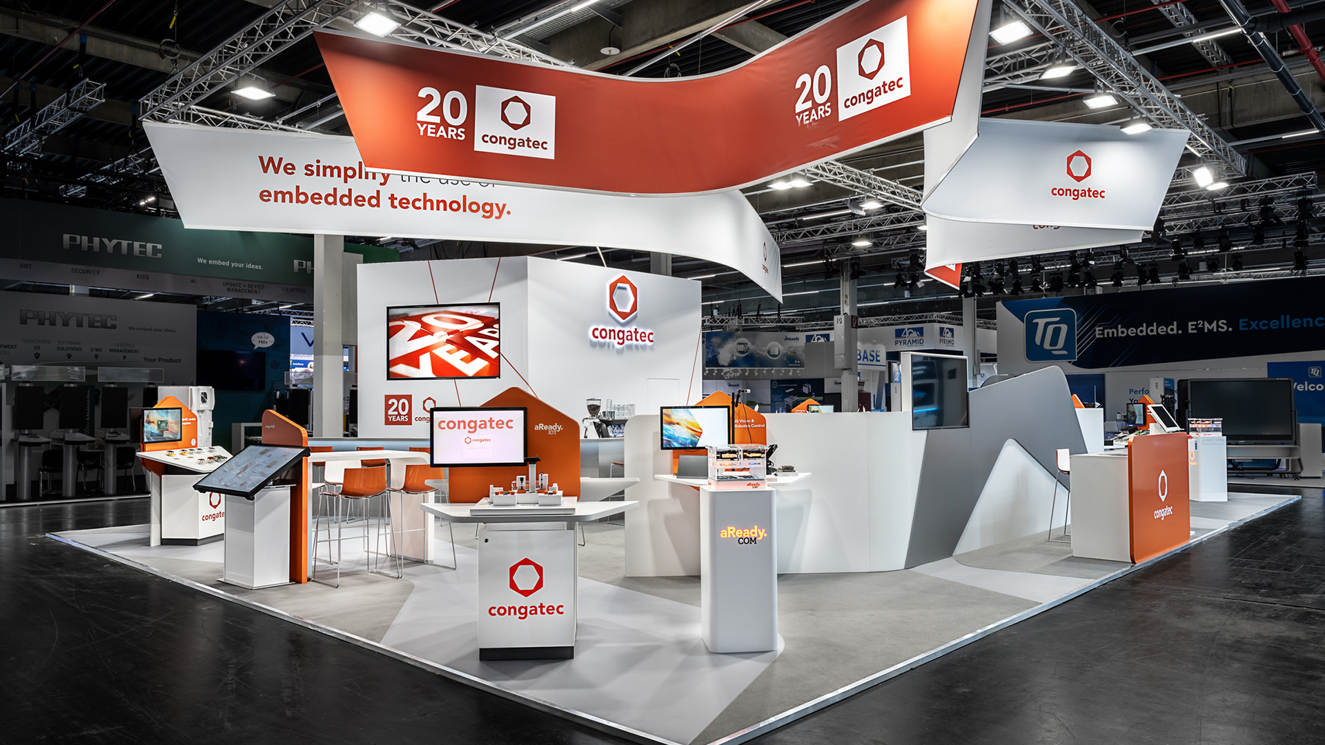 Messestand von Congatec auf der embedded world in Nürnberg mit 170 m² Ausstellungsfläche. Die geschwungenen Deckenbanner in Weiß und Orange greifen die Formensprache des Congatec-Logos auf und schaffen eine starke Fernwirkung. Organisch geformte Wände, wiederverwendbare Produktpräsenter, interaktive Touchscreens und hochwertige Präsentationsflächen inszenieren Embedded-Computing-Lösungen in einem modernen Markenauftritt. Das klare Farbkonzept in Orange und Weiß, kombiniert mit fließenden Formen und nachhaltigen Standbauelementen, sorgt für eine hohe Wiedererkennbarkeit und eine offene, einladende Messearchitektur.