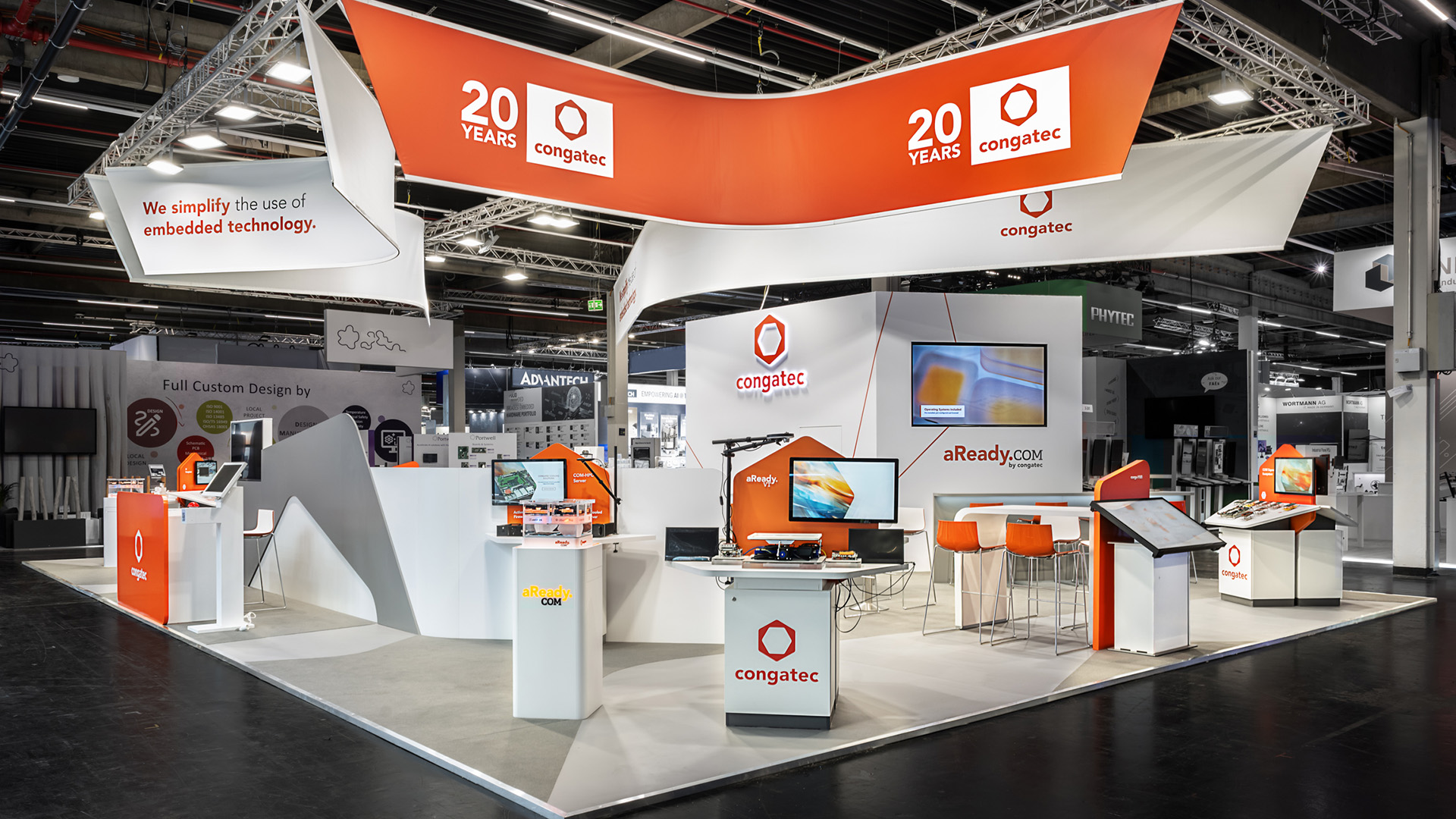 Messestand von Congatec auf der embedded world in Nürnberg mit 170 m² Ausstellungsfläche. Die markanten, geschwungenen Deckenbanner in der Corporate-Farbe Orange verleihen dem Stand eine hohe Fernwirkung und greifen die Formensprache des Congatec-Logos auf. Organisch gestaltete Wände, modulare Produktpräsenter, interaktive Touchscreens sowie moderne Besprechungsbereiche schaffen eine offene und einladende Präsentationsfläche für Embedded-Computing-Technologien. Nachhaltig konzipierte Standbauelemente, hochwertige Materialien und die konsequente Umsetzung der Corporate Identity sorgen für einen innovativen Messeauftritt mit hohem Wiedererkennungswert.