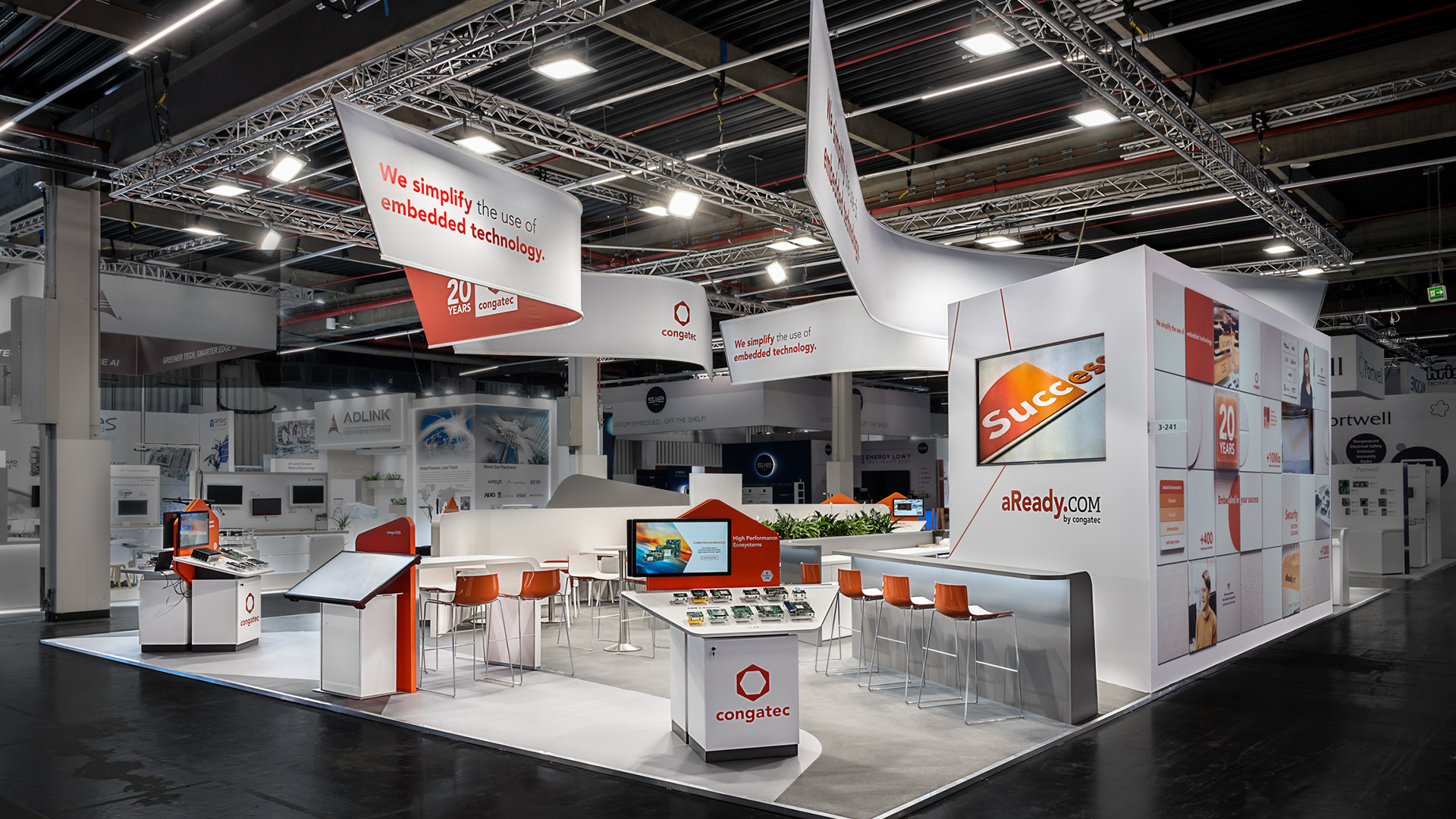 Messestand von Congatec auf der embedded world in Nürnberg mit 170 m² Ausstellungsfläche. Der offene Markenauftritt verbindet geschwungene Deckenbanner mit organisch geformten Architektur-Elementen und greift konsequent die abgerundete Formensprache des Congatec-Logos auf. Die auffällige Corporate-Farbe Orange setzt gezielte Akzente und führt Besucher zu interaktiven Produktpräsentationen, Touchscreens und modernen Kommunikationsbereichen. Modulare Präsentationsmöbel, nachhaltige Standbauelemente und eine klare Besucherführung schaffen einen hochwertigen Messeauftritt, der Embedded-Computing-Technologien, Innovation und Markenidentität eindrucksvoll in Szene setzt.
