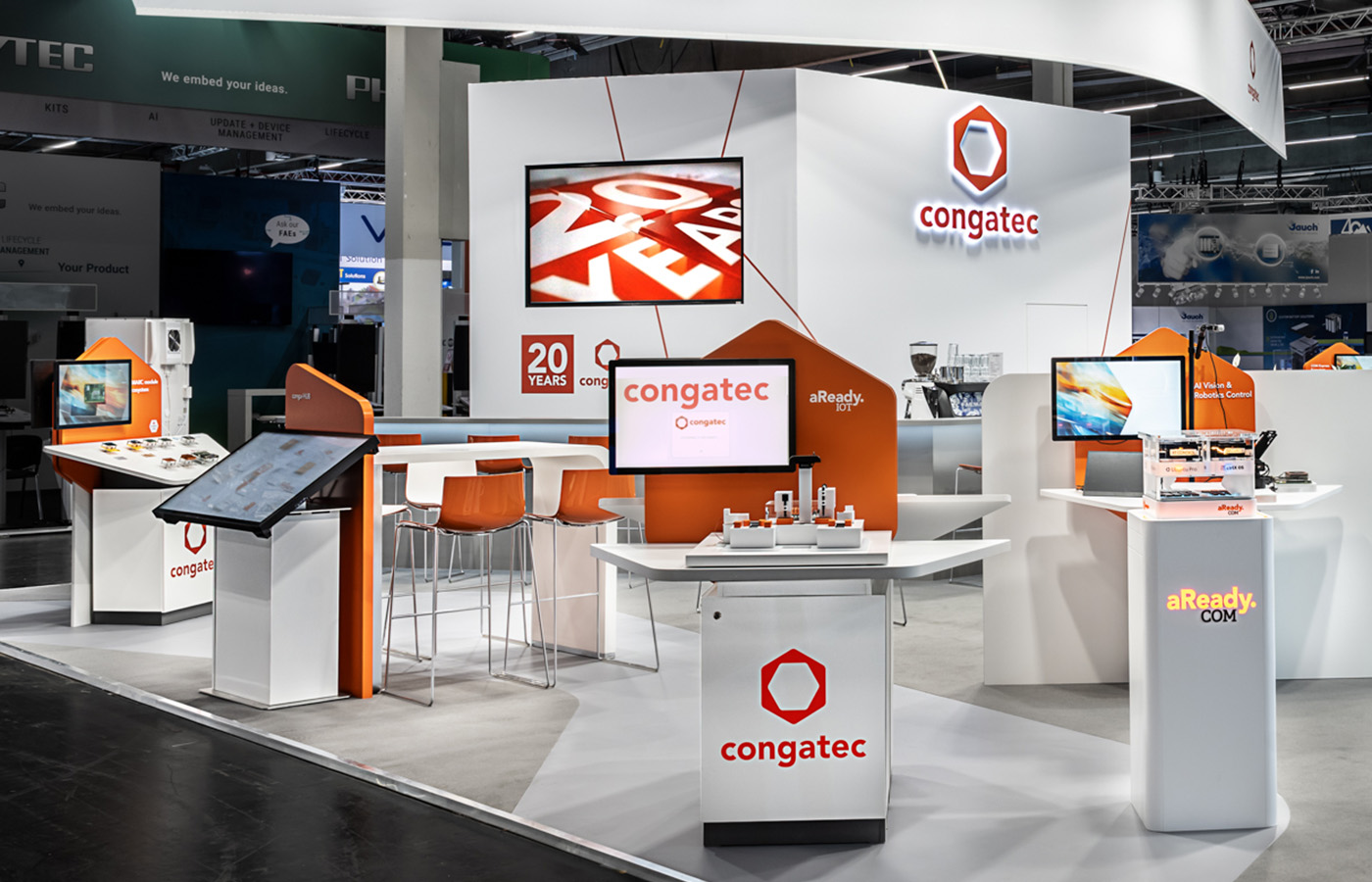 Detailansicht des Congatec-Messestands auf der embedded world in Nürnberg mit interaktiven Produktpräsentationen für Embedded-Computing-Technologien. Im Mittelpunkt stehen modular entwickelte Produktpräsenter, Touchscreen-Terminals und digitale Demonstrationsflächen in der markanten Corporate-Farbe Orange. Die organisch geformten Möbel und Wandelemente greifen die Formensprache des Congatec-Logos auf und schaffen gemeinsam mit den offenen Kommunikationsbereichen eine moderne, einladende Messearchitektur. Das nachhaltige Standkonzept kombiniert hochwertige Materialien, wiederverwendbare Präsentationssysteme und eine klare Markeninszenierung für maximale Aufmerksamkeit auf internationalen Technologiemessen.