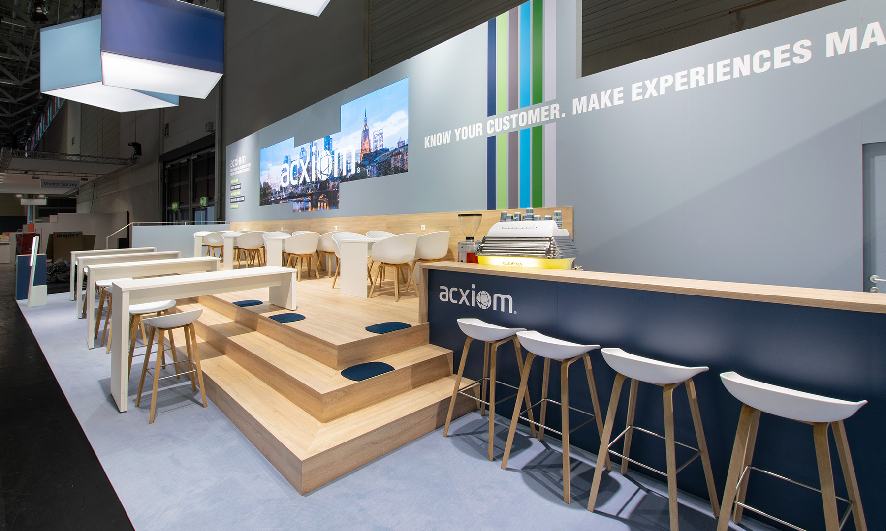 Detailaufnahme des Acxiom-Messestands auf der dmexco in Köln. Das großzügige Treppenpodest aus hellem Holz bildet das architektonische Herzstück des 75 m² großen Messestands und verbindet verschiedene Kommunikationsbereiche miteinander. Im Hintergrund präsentieren großformatige LED-Screens dynamische Markeninhalte und sorgen gemeinsam mit der grafisch gestalteten Rückwand für eine aufmerksamkeitsstarke Inszenierung. Die Kombination aus natürlichen Holzoberflächen, der blaugrauen Corporate Identity, offenen Sitzbereichen und einer integrierten Bar schafft eine einladende Atmosphäre für Beratung, Networking und Kundengespräche. Abgehängte Logoquader erhöhen die Fernwirkung und machen den Markenauftritt bereits aus großer Entfernung sichtbar.