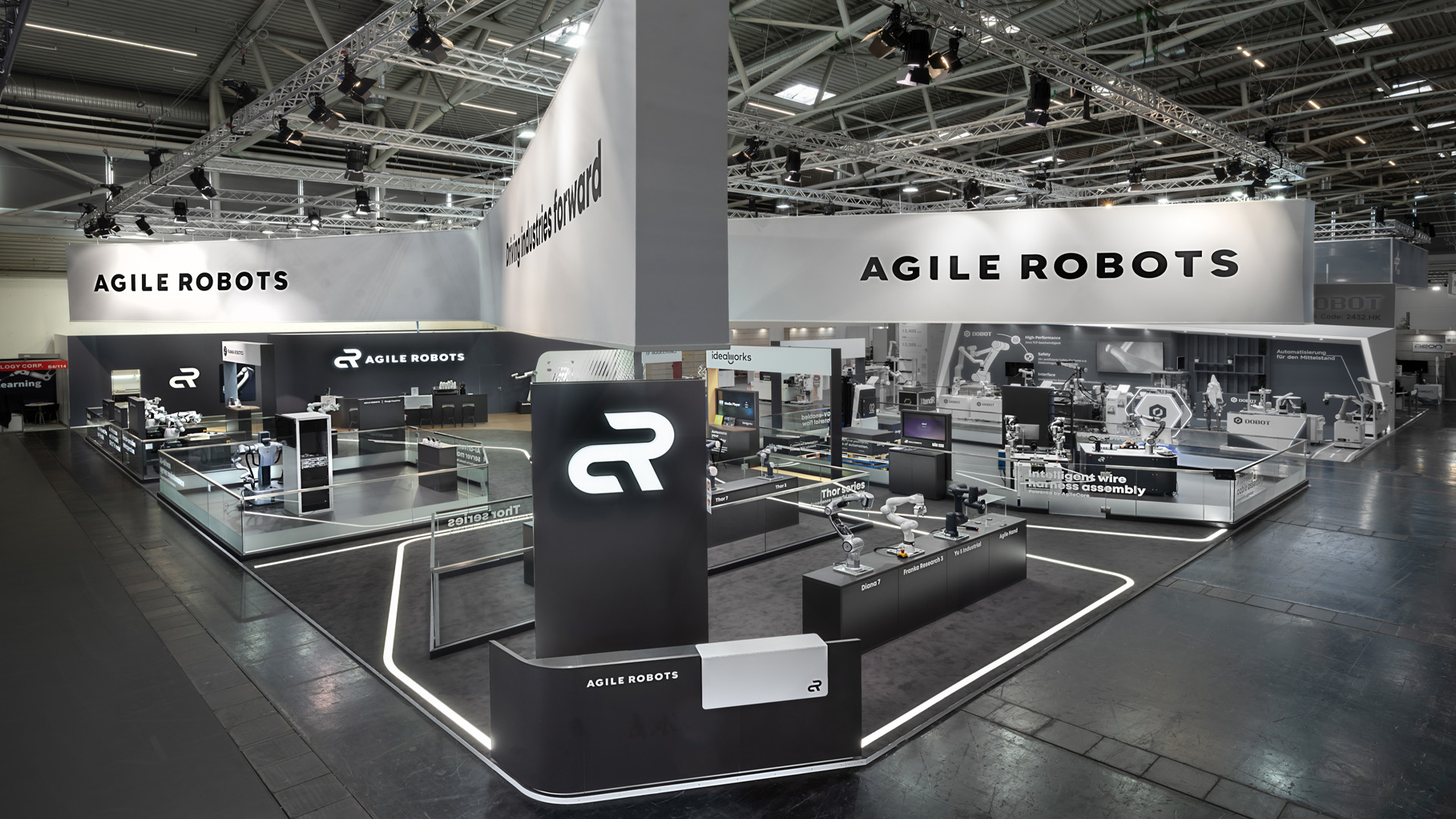 Panoramaansicht des 515 m² großen Messestands von Agile Robots auf der automatica in München. Der markante kreuzförmige Deckenbanner mit dem Unternehmenslogo verbindet die offenen Präsentationsflächen, während LED-Lichtlinien Besucher durch die Ausstellung mit Industrie-Robotern, KI- und Automatisierungslösungen führen.
