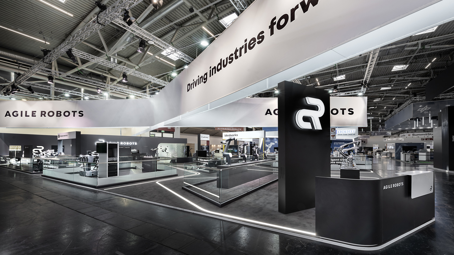 Großzügiger Messestand von Agile Robots auf der automatica in München mit 515 m² Ausstellungsfläche. Der markante kreuzförmige Deckenbanner mit dem Claim „Driving industries forward“, beleuchtete Bodenlinien und offene Präsentationsflächen schaffen eine moderne Bühne für Industrie-Robotik, KI und Automatisierungslösungen.