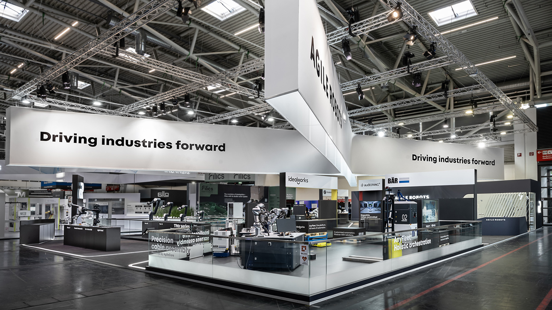 Gesamtansicht des 515 m² großen Messestands von Agile Robots auf der automatica in München. Der markante kreuzförmige Deckenbanner verbindet die Präsentationsbereiche der Tochterunternehmen, während klare Architektur, reduzierte Materialien und beleuchtete Wegeführung eine moderne Bühne für Industrie-Roboter und Automatisierungslösungen schaffen.