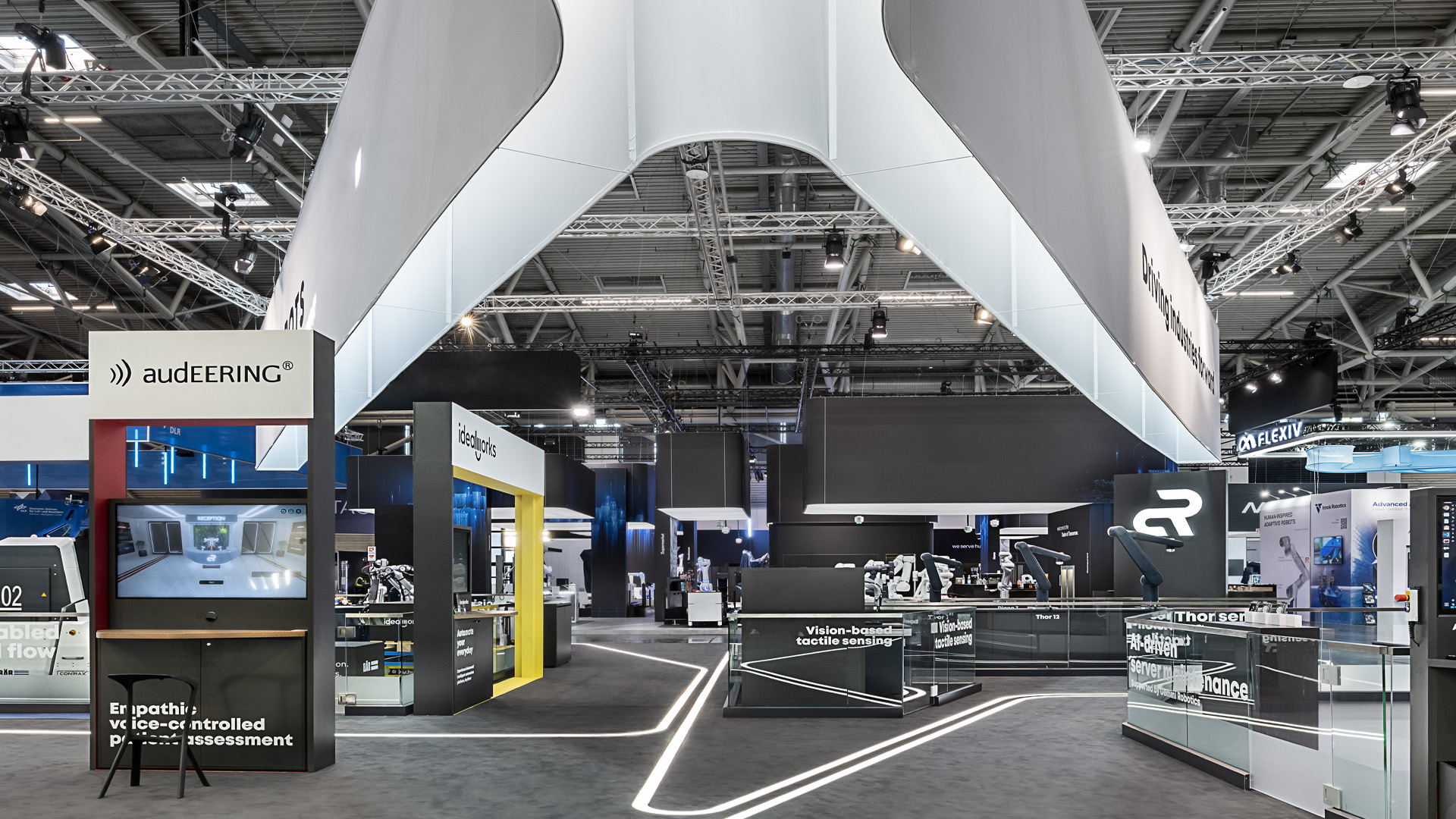 Großzügiger Messestand von Agile Robots auf der automatica in München mit 515 m² Ausstellungsfläche. Der überdimensionale, kreuzförmige Deckenbanner verbindet die Präsentationsbereiche der Tochterunternehmen, während integrierte Leuchtstreifen Besucher intuitiv durch die moderne Robotik- und Automatisierungsausstellung führen.