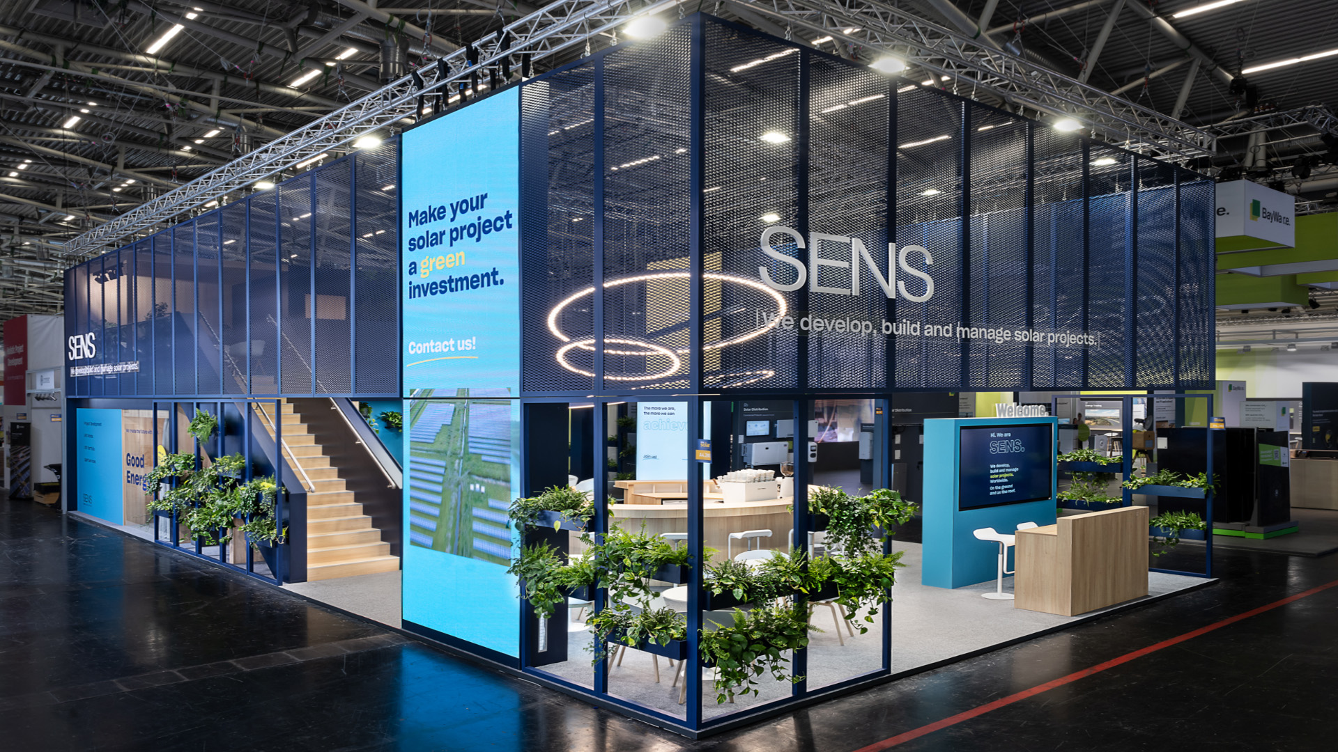 Außenansicht des zweigeschossigen SENS Messestands auf der Intersolar Smarter E in München mit markanter Streckmetall-Fassade, großformatigen LED-Medienflächen, offenem Eingangsbereich, begrünten Fassadenelementen und zentraler Kommunikationszone. Das 192 m² große Messekonzept vereint moderne Architektur, hohe Fernwirkung und nachhaltiges Design und schafft vielfältige Bereiche für Beratung, Networking und Produktpräsentationen.