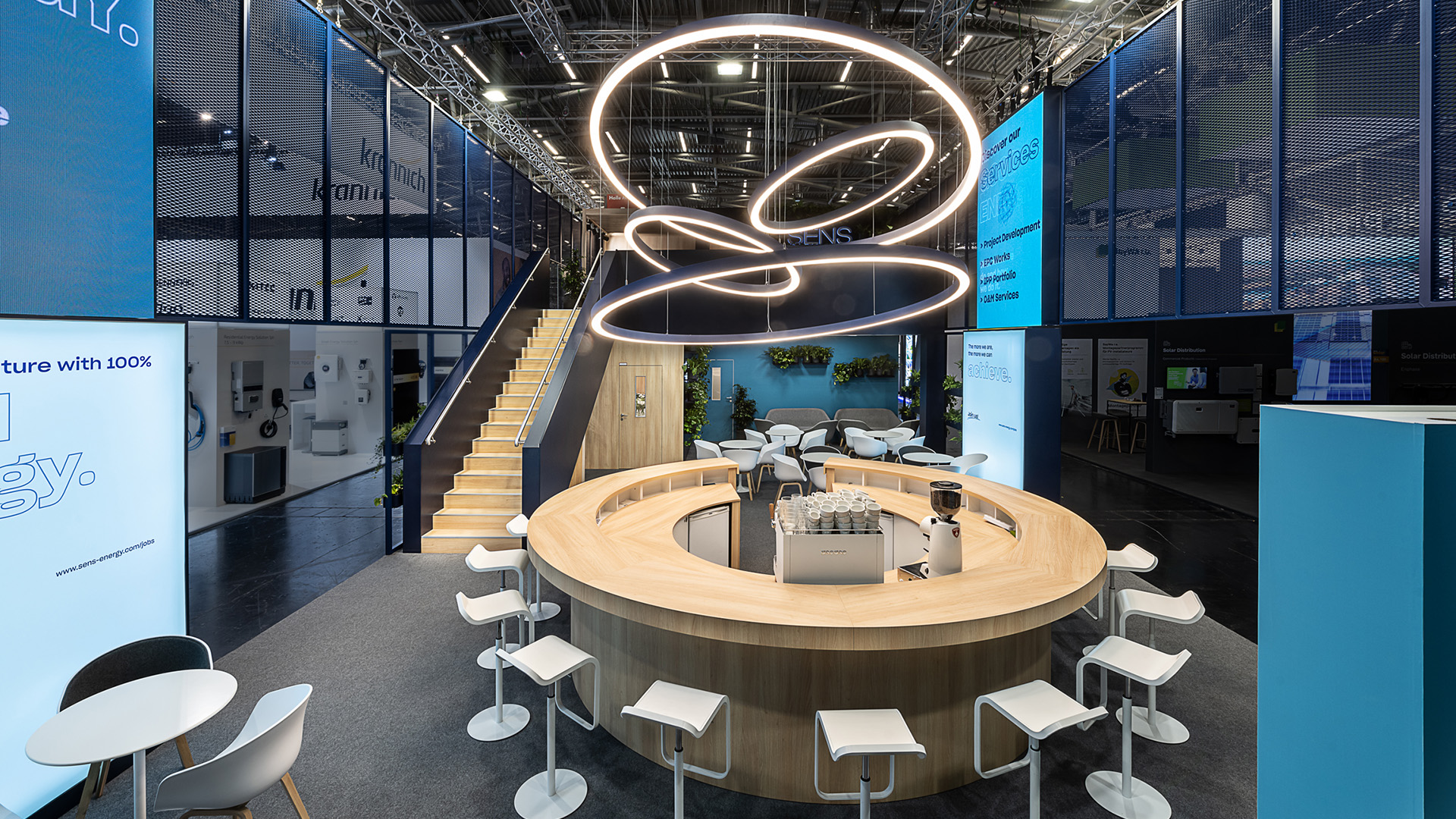 Innenansicht des zweigeschossigen SENS Messestands auf der Intersolar Smarter E in München mit zentraler runder Kommunikationsbar, skulpturalem LED-Lichtobjekt, transparenter Streckmetall-Fassade, Treppenaufgang zum Obergeschoss und begrünten Loungebereichen. Das offene Standkonzept fördert Networking, Beratung und persönliche Gespräche auf insgesamt 192 m² Ausstellungsfläche.