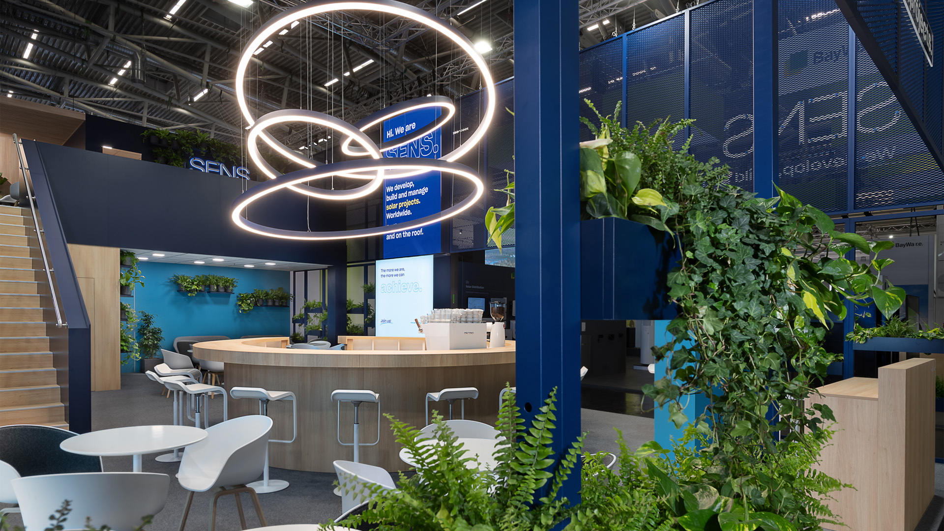 Perspektivische Innenansicht des zweigeschossigen SENS Messestands auf der Intersolar Smarter E in München mit runder Kommunikationsbar, skulpturalem LED-Lichtobjekt, begrünten Pflanzenelementen, transparenter Streckmetall-Fassade und offenen Meetingbereichen. Das nachhaltige Standkonzept verbindet moderne Architektur, natürliche Materialien und vielfältige Kommunikationszonen auf einer 192 m² großen Ausstellungsfläche.