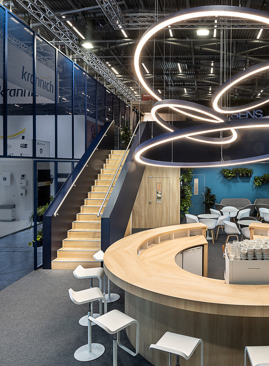 A detailed view of the SENS exhibition booth at Intersolar Smarter E in Munich, featuring a central circular communication bar, a sculptural LED light installation, an open staircase leading to the second level, a transparent expanded metal frame, and green lounge areas. The modern booth architecture combines open communication, sustainable design, and a high-quality environment for consultations and networking. Translated with DeepL.com (free version)