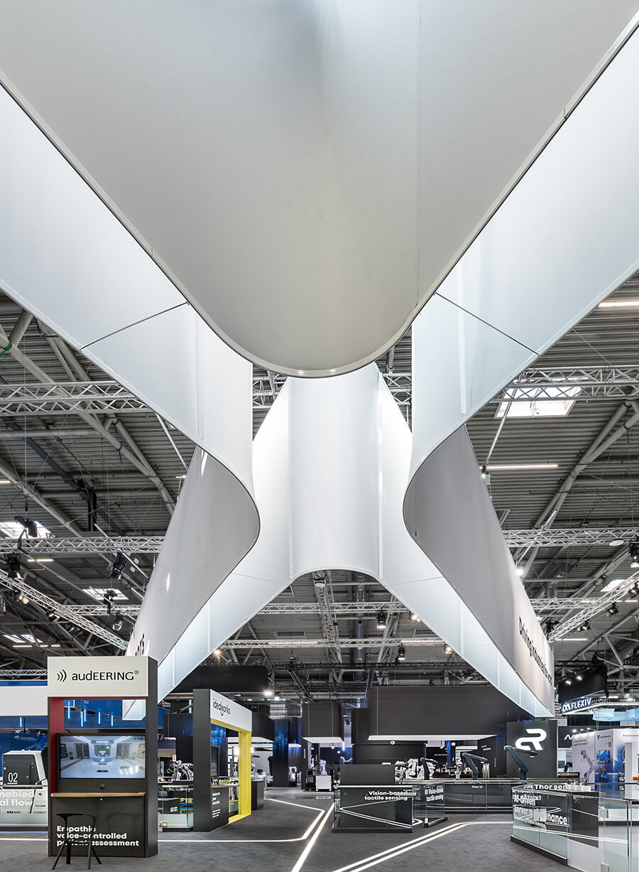 Messestand von Agile Robots auf der automatica in München mit markantem kreuzförmigem Deckenbanner, offenem Standkonzept, klaren Sichtachsen, integrierten LED-Leitlinien und reduzierter Architektur zur Präsentation moderner Industrieroboter und Automatisierungslösungen.