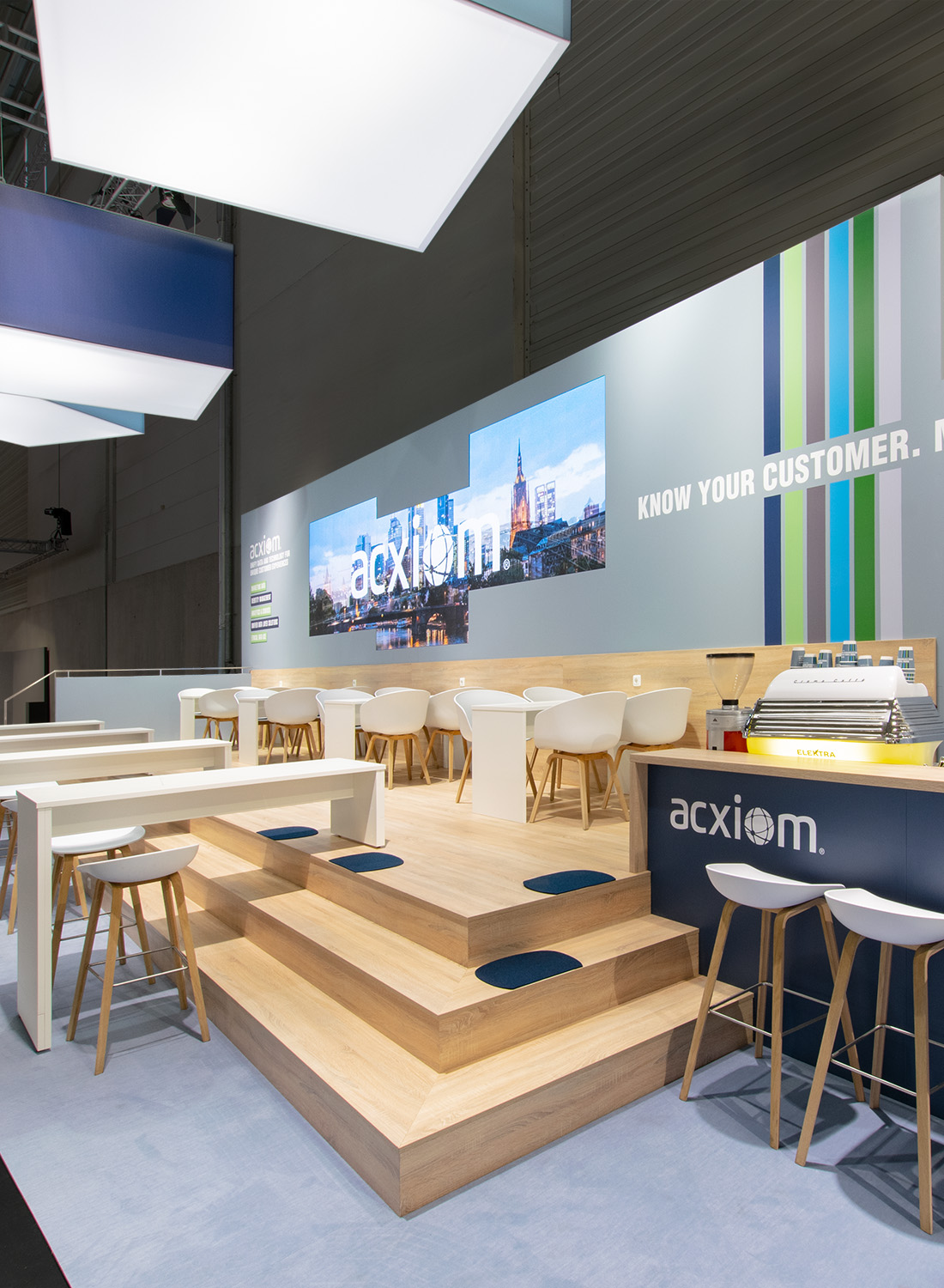 Close-up of the Acxiom booth at dmexco in Cologne. The spacious staircase landing made of light-colored wood forms the architectural centerpiece of the 75 m² booth and connects various communication areas. In the background, large-format LED screens display dynamic brand content and, together with the graphically designed back wall, create an eye-catching presentation. The combination of natural wood surfaces, the blue-gray corporate identity, open seating areas, and an integrated bar creates a welcoming atmosphere for consultations, networking, and client meetings. Suspended logo cubes enhance the stand’s visibility from a distance, making the brand presence visible even from far away.