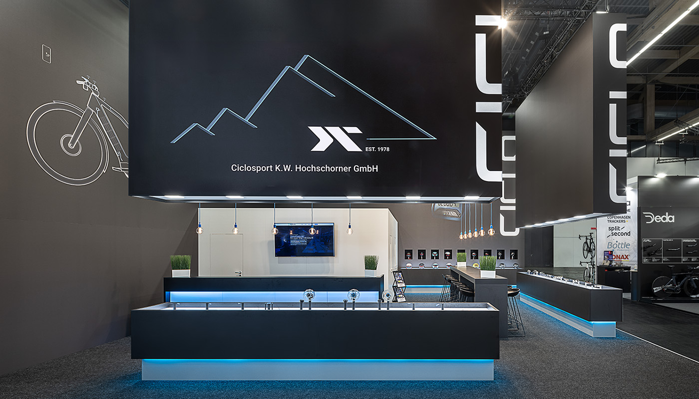 Ciclo Sport e-Bike Components’ modern booth at Eurobike in Frankfurt featured oversized black ceiling cubes, illuminated product display cases, a contrasting white booth structure, and subtle blue branding accents for an eye-catching product presentation.