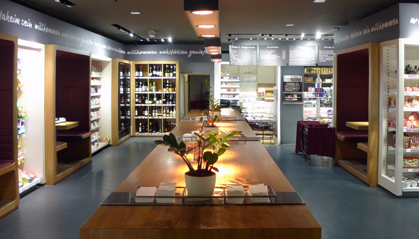 Zentraler Gemeinschaftstisch im Kramer's Café- und Convenience Store in Salzburg und Innsbruck mit integrierten Sitznischen, Weinregal und Gastronomiebereich.
