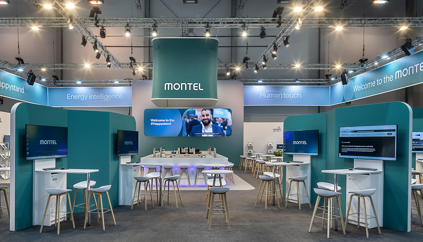 A symmetrical interior view of the Montel exhibition booth at E-world in Essen, featuring a central hospitality bar, a suspended brand cube, digital information stations, open meeting areas, and modern booth architecture in the company’s green corporate identity.