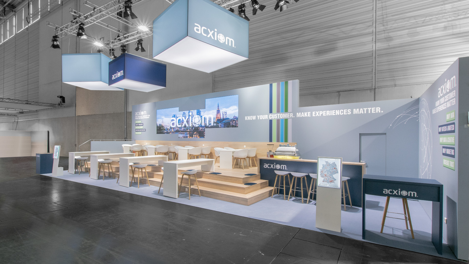 Gesamtansicht des Acxiom Messestandes auf der DMEXCO in Köln mit beleuchteten Logoquadern, großformatiger LED-Medienwand, erhöhtem Holzpodest und offenen Kommunikationsbereichen für Kundengespräche und Networking.