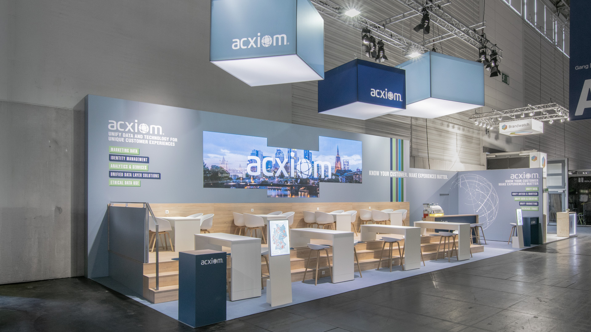 Gesamtansicht des Acxiom Messestandes auf der DMEXCO in Köln mit beleuchteten Logoquadern, großformatiger LED-Medienwand, erhöhtem Holzpodest und offenen Kommunikationsbereichen für Beratung und Networking.