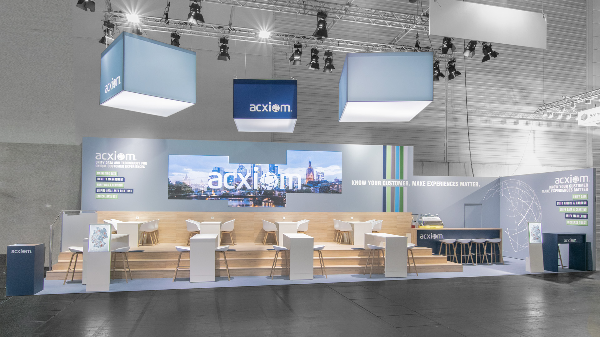 Individuell gestalteter Acxiom Messestand auf der DMEXCO in Köln mit abgehängten Logoquadern, LED-Wand, Kommunikationspodest und moderner Messearchitektur.