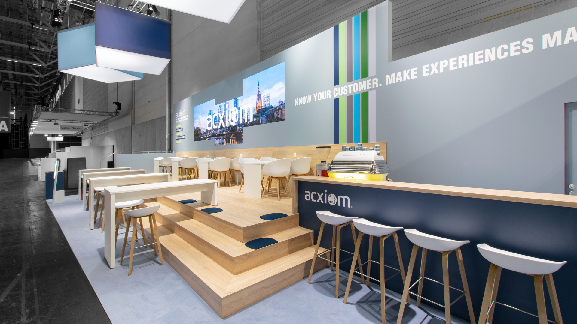 Kommunikationsbereich des Acxiom Messestandes auf der DMEXCO in Köln mit erhöhtem Holzpodest, Bar, LED-Medienwand und beleuchteten Logoquadern für moderne Markenkommunikation.