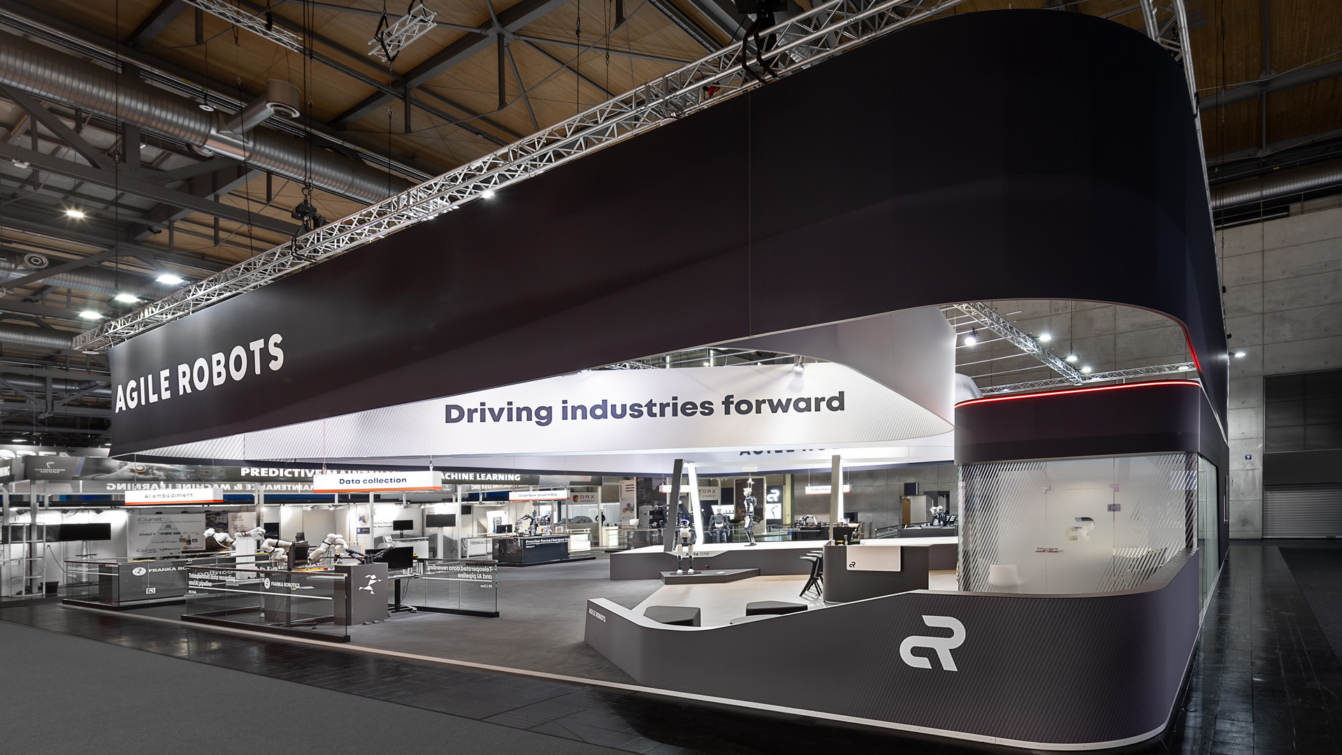 Großflächiger Messestand von Agile Robots auf der Hannover Messe mit markantem, umlaufendem Deckenbanner, offener Standarchitektur und modernen Demonstrationsflächen für industrielle Robotik. Die klare Gestaltung, hochwertige Materialien und präzise Lichtführung schaffen eine starke Markenpräsenz und setzen innovative Automatisierungs- und KI-Lösungen wirkungsvoll in Szene.