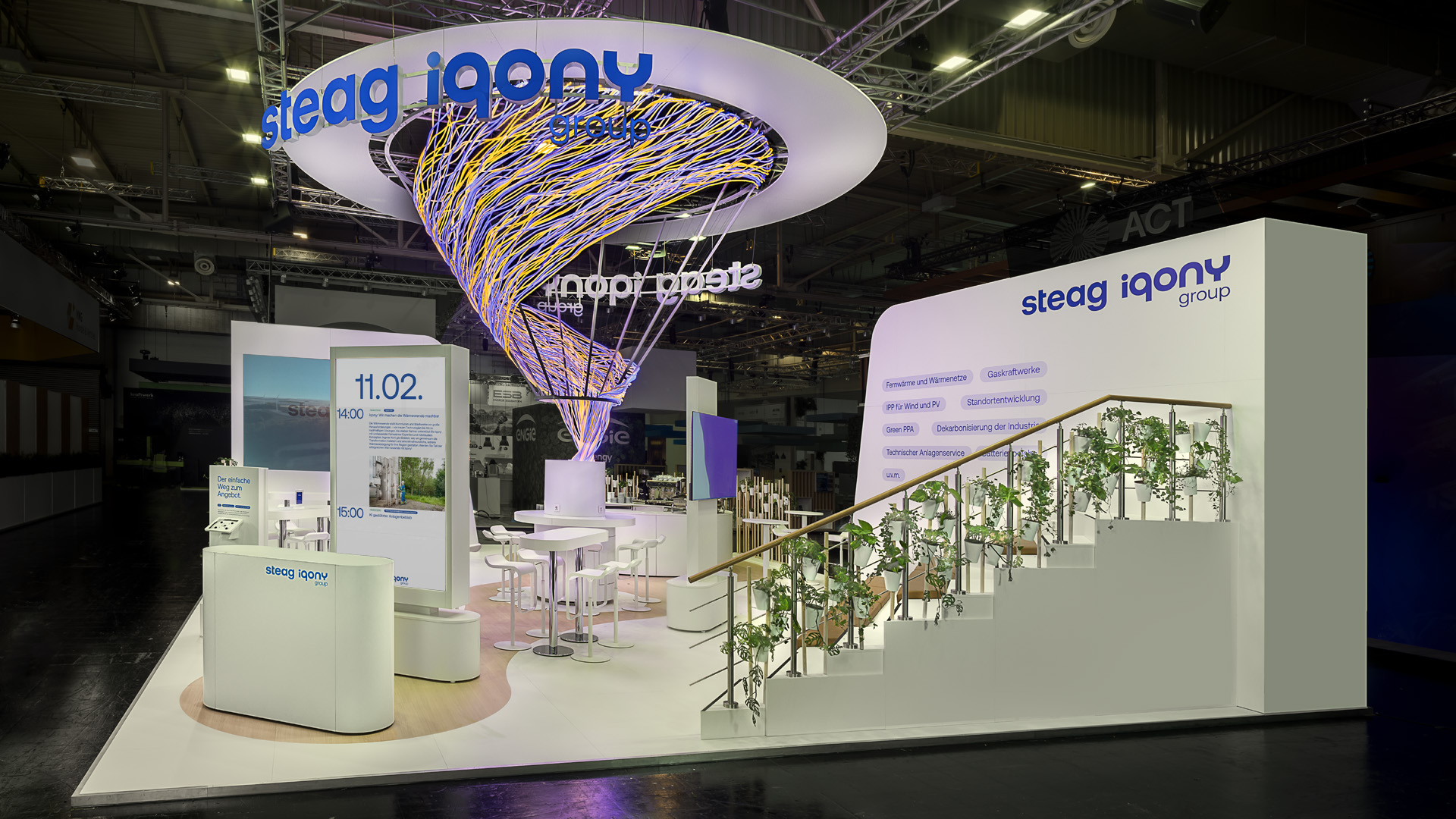 Perspektivischer Blick auf den Messestand der STEAG Iqony Group auf der E-world mit einer spektakulären, schwebenden Lichtskulptur als zentralem Markenzeichen. Die offene Architektur, begrünte Treppenanlage und digitale Informationsflächen schaffen eine einladende Atmosphäre für Begegnung, Beratung und den Austausch zu nachhaltigen Energielösungen.