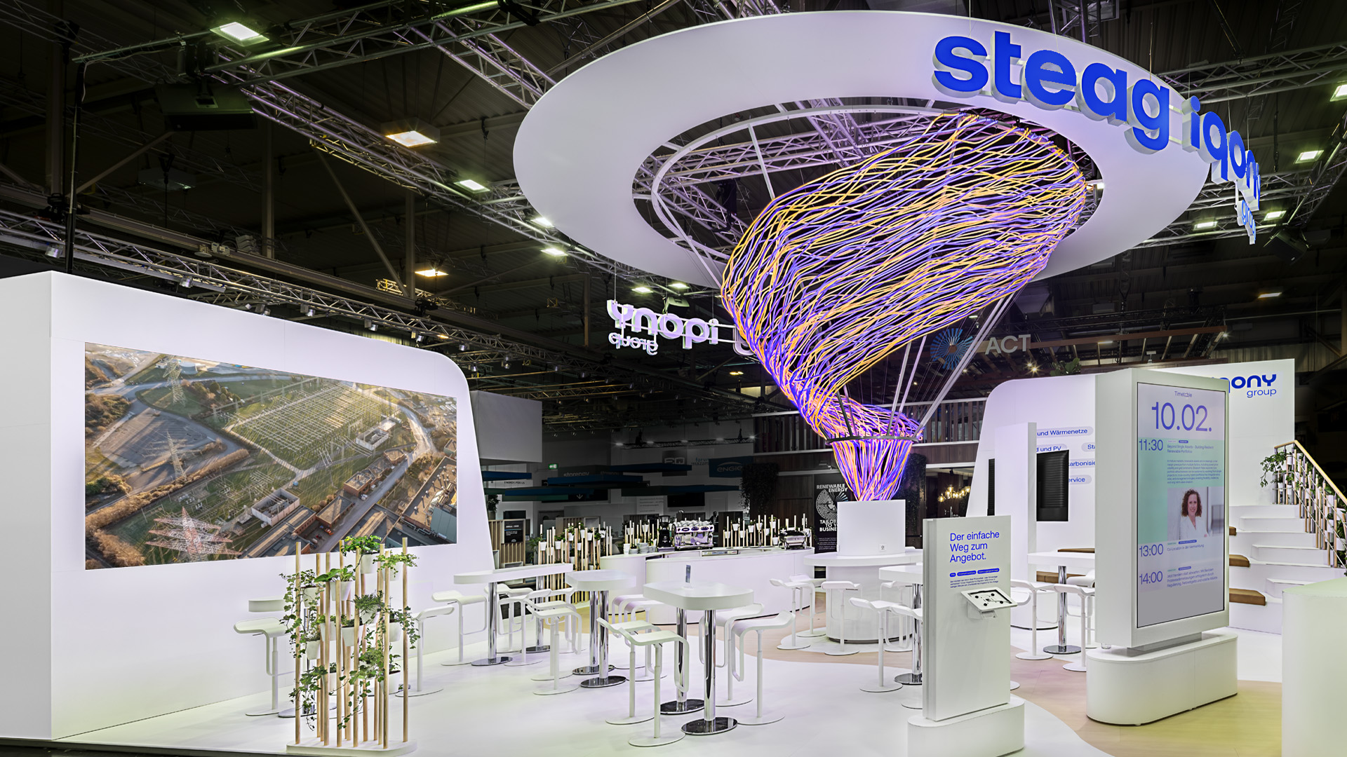 Perspektivischer Blick in den Messestand der STEAG Iqony Group auf der E-world mit einer spektakulären, dynamischen Lichtskulptur als zentralem Gestaltungselement. Die offene Standarchitektur, großzügige Kommunikationszonen und digitale Präsentationsflächen schaffen eine hochwertige Umgebung für Beratung, Wissensaustausch und Markeninszenierung.