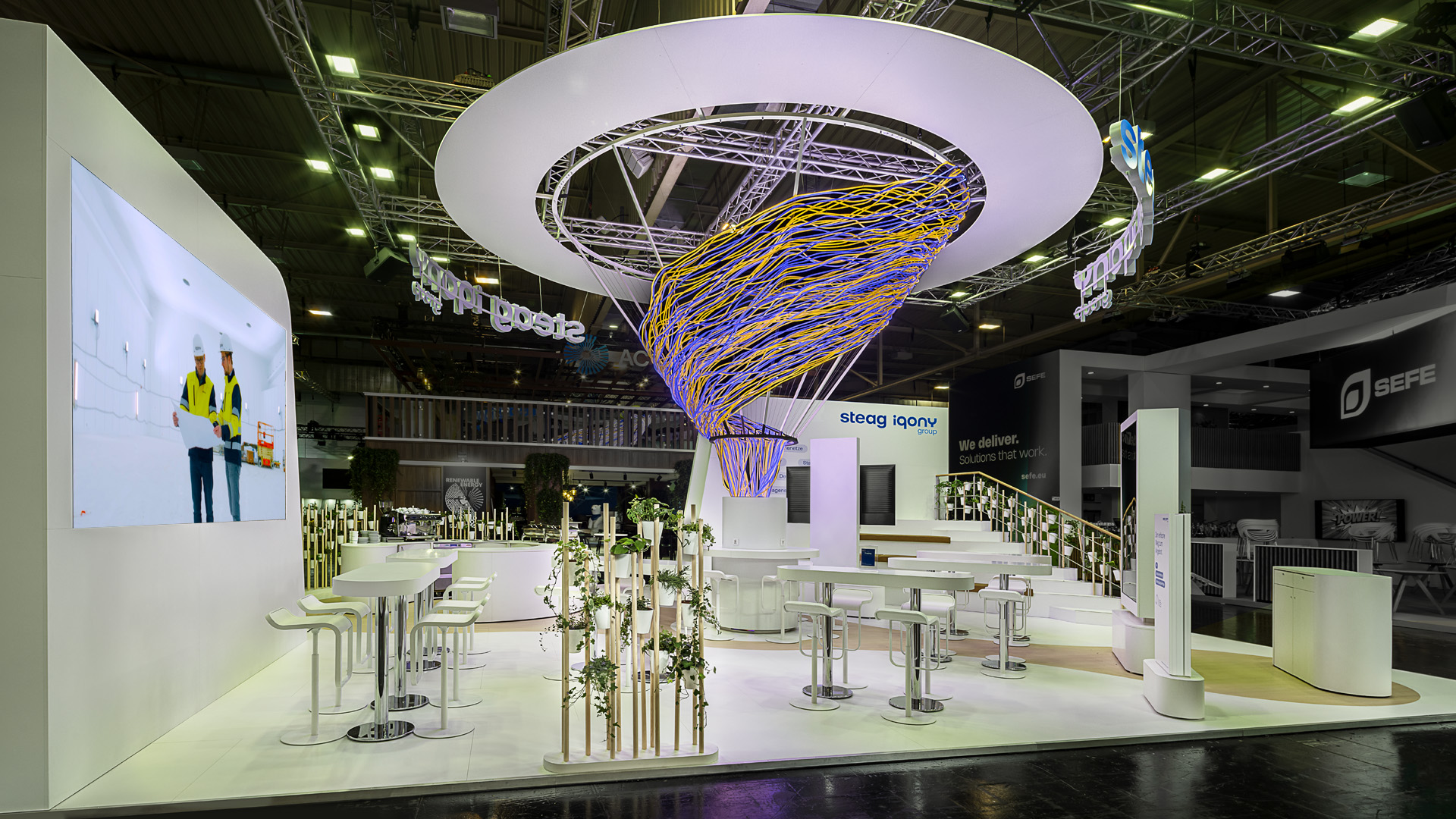 Messestand der STEAG Iqony Group auf der E-world mit einer spektakulären Lichtskulptur als architektonischem Mittelpunkt. Offene Kommunikationsbereiche, großzügige Präsentationsflächen und begrünte Gestaltungselemente schaffen eine einladende Atmosphäre und verbinden Innovation, Nachhaltigkeit und persönliche Begegnung zu einem modernen Markenauftritt.
