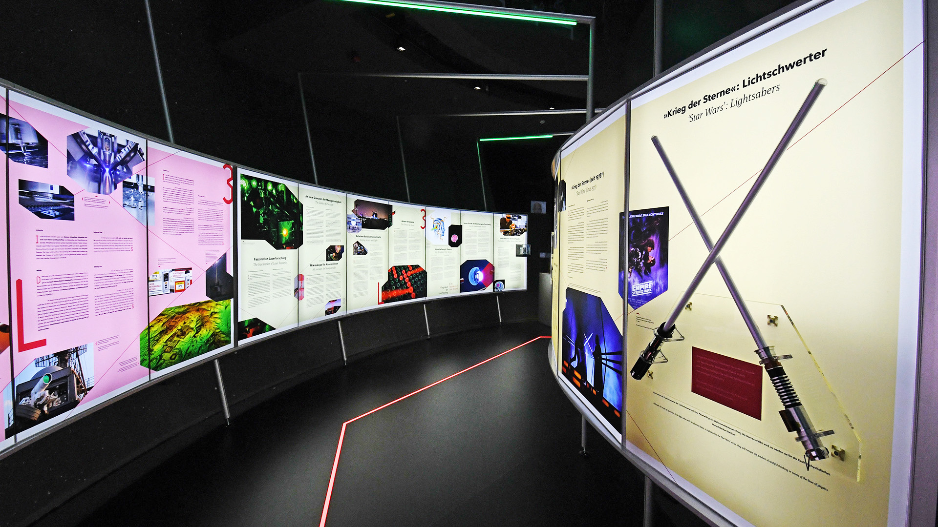 Ausstellungsstation der Sonderausstellung „Laser Licht Leben“ im ESO Planetarium Garching mit gebogenen Informationstafeln, Exponat zu Star-Wars-Lichtschwertern, interaktiver Wissensvermittlung und moderner Lichtinszenierung rund um Lasertechnologie.