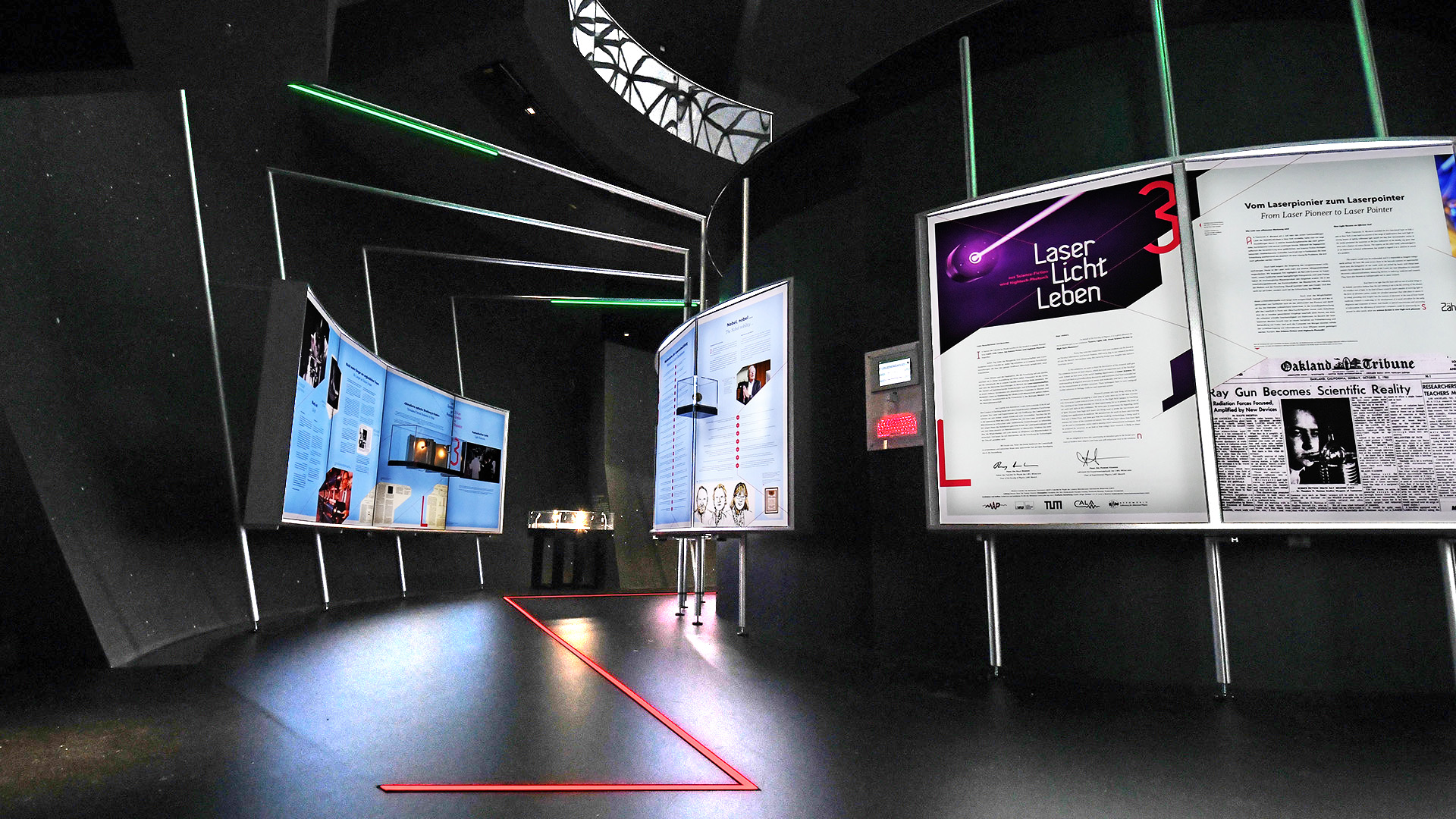 Interaktiver Ausstellungsbereich der Sonderausstellung „Laser Licht Leben“ im ESO Planetarium Garching mit beleuchteten Informationstafeln, Exponaten, LED-Lichtlinien und immersiver Szenografie zur Geschichte und Anwendung moderner Lasertechnologie.