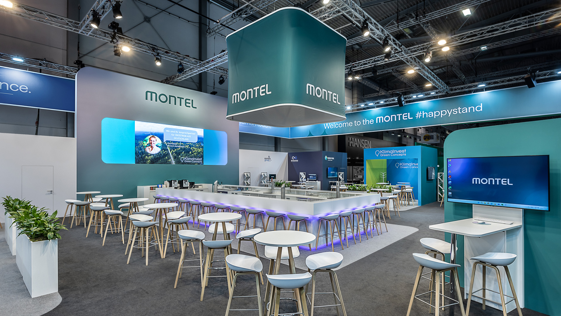 Zentraler Hospitality-Bereich des Montel Messestands auf der E-world in Essen mit großzügiger Bar, offenen Sitzbereichen, digitalen Präsentationsflächen, markantem Deckenkubus und moderner Standarchitektur in der grünen Corporate Identity für Networking und Fachgespräche.