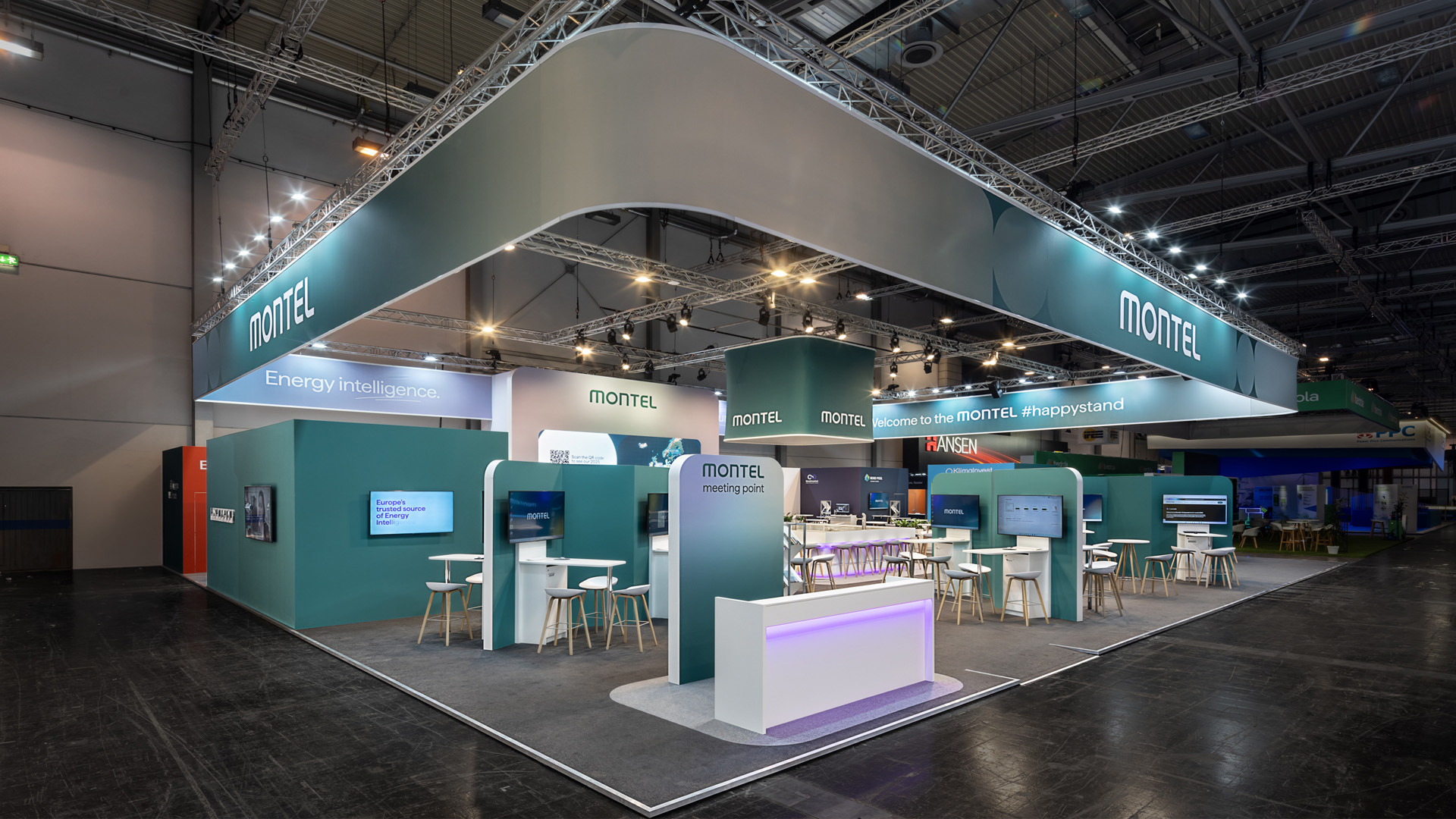 Offener Messestand von Montel auf der E-world in Essen mit großem umlaufenden Deckenbanner, einladendem Meeting Point, digitalen Präsentationsstationen, großzügigen Kommunikationsbereichen und moderner Architektur in der grünen Corporate Identity des Energie-Informationsdienstleisters.