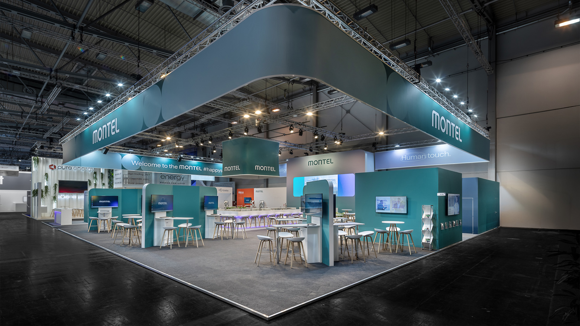 Großzügiger Messestand für Montel auf der E-world in Essen mit markantem, umlaufendem Deckenbanner, organisch gerundeter Architektur, offenen Kommunikationsbereichen, digitalen Präsentationsflächen und moderner Standgestaltung in der charakteristischen grünen Corporate Identity.