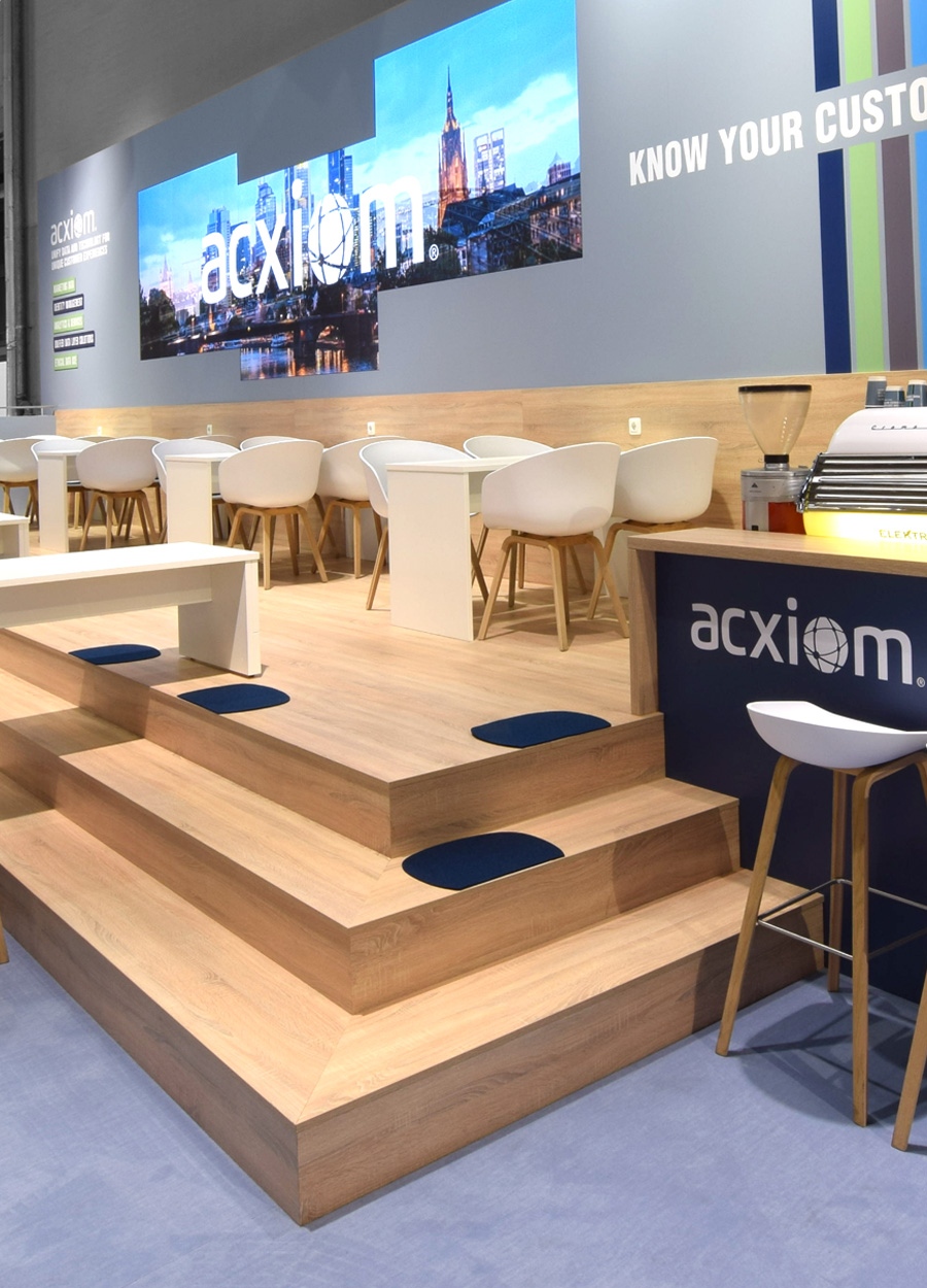 Moderner Acxiom Messestand auf der dmexco in Köln mit markantem Holzpodest als Kommunikationsplattform, großzügigen Sitzbereichen, großformatiger LED-Medienwand, hochwertiger Hospitality-Zone und zeitgemäßer Standarchitektur in der Corporate Identity des Unternehmens.