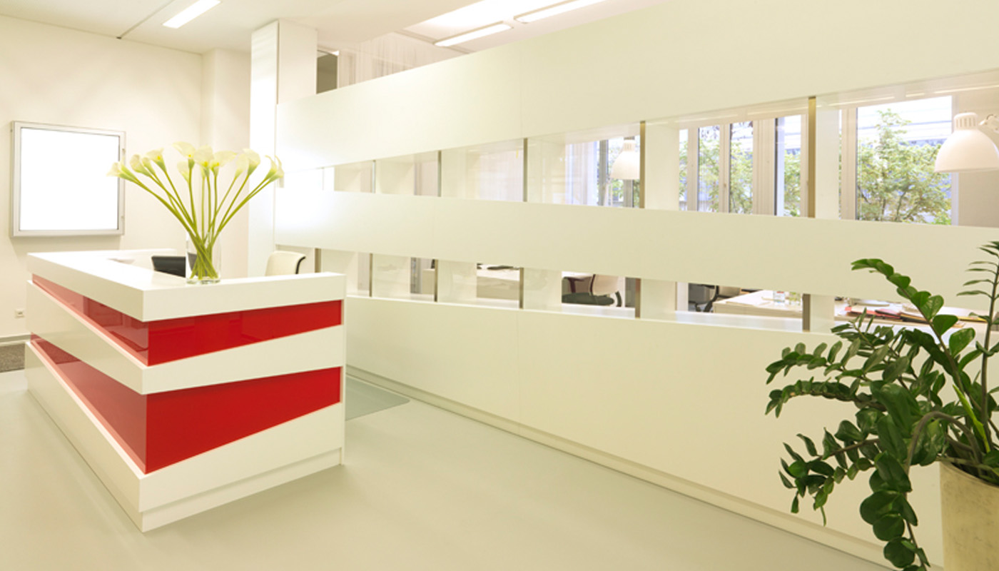 Moderne Rezeption einer Werbeagentur in München mit weißem Empfangstresen, roten Designelementen und offenem Raumteiler mit horizontalen Sichtachsen.