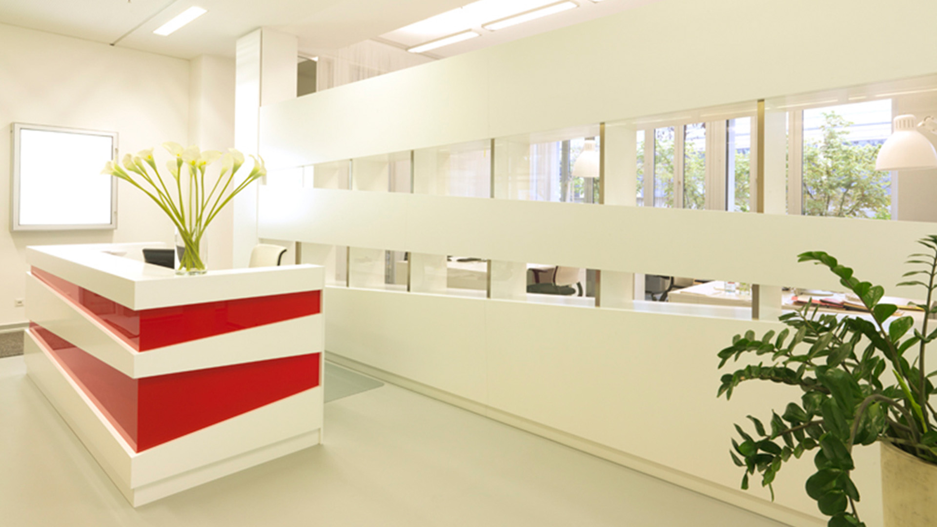 Moderne Rezeption einer Werbeagentur in München mit weißem Empfangstresen, roten Designelementen und offenem Raumteiler mit horizontalen Sichtachsen.