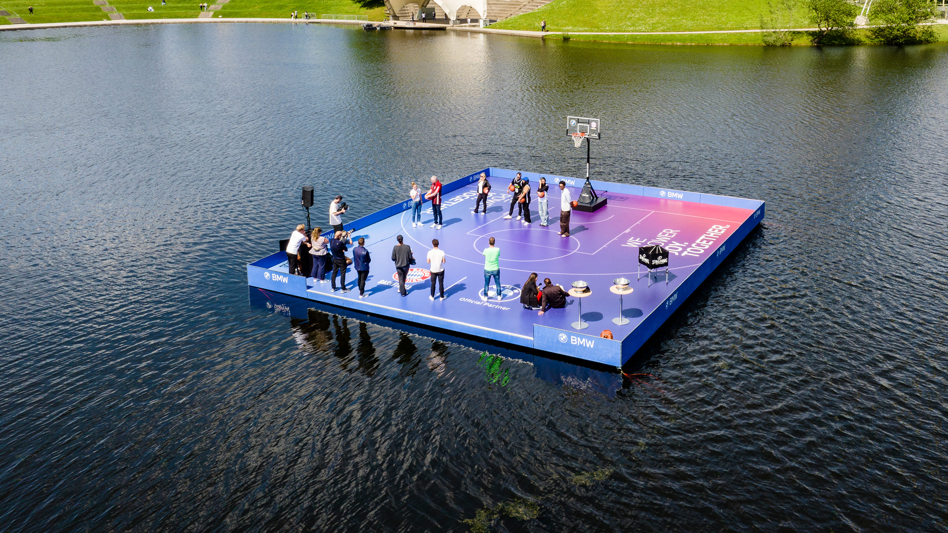 Schwimmendes Basketballfeld auf dem Olympiasee in München während eines Events für BMW und den FC Bayern Basketball mit Zuschauern und Basketballkorb auf einer schwimmenden Plattform.
