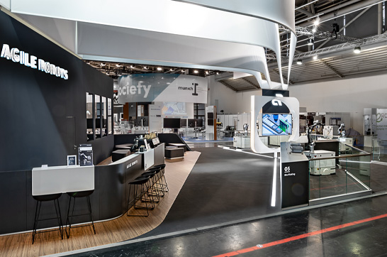 Offener Messestand von Agile Robots auf der automatica in München mit geschwungenem Deckenbanner, großzügigem Hospitality-Bereich, moderner Produktpräsentation und klarer Architektur für Robotik- und Automatisierungslösungen.